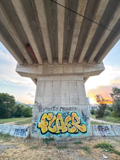 The image shows the underside of a concrete overpass with several parallel beams. A red pipe runs along the underside of one beam. The concrete pillar supporting the overpass has graffiti on it, including a large yellow tag with blue outlines and black lettering above it. Other smaller graffiti tags are visible on the pillar and on concrete barriers in the foreground. The ground is covered in dry grass and some litter. Trees and buildings are visible in the background, with a sunset sky.