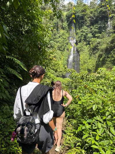 Two individuals, viewed from behind, are walking on a path towards a waterfall in a lush, green jungle. The person in the foreground wears a white shirt, dark shorts, and a black backpack. A black jacket is draped over their shoulders. The second individual, slightly ahead, wears a black swimsuit and has wet hair. The waterfall cascades down a rocky, vegetation-covered cliff face. Palm trees are visible in the background, suggesting a tropical environment. The scene is located in the city of Sawan, Indonesia.