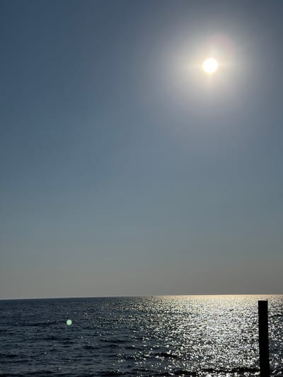 The image depicts a serene seascape under a clear sky, likely captured in Buljarica, Montenegro. The sun, a bright orb with visible rays, is positioned in the upper right quadrant, casting a dazzling sheen across the water's surface. The sea itself is a deep blue, with ripples and waves reflecting the sunlight, creating a shimmering, almost glittery effect. In the foreground, on the right side of the frame, a dark, vertical post, possibly part of a pier or jetty, stands in silhouette against the bright water. The horizon line is sharp, separating the vast expanse of the sky from the sea. There are no visible people or explicit human activities in the scene, though distant, faint specks on the water might indicate small boats. The overall atmosphere is one of tranquility and natural beauty, characteristic of a coastal environment.