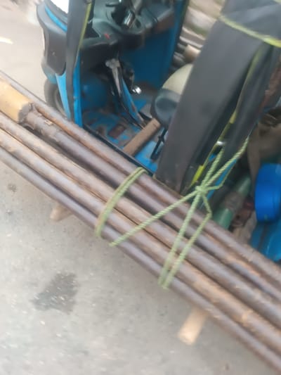 A bundle of brown metal pipes, secured with green twine, rests on a gray surface, likely a road or pavement. In the background, a blue motorized tricycle is partially visible, with parts of its cabin and seat showing. The overall impression is that the pipes are being transported by the tricycle, possibly for construction or other utility purposes.