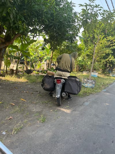 In Karangtanjung, Indonesia, a person wearing a green hooded sweatshirt and light-colored pants is seated on a motorcycle. The motorcycle is equipped with two black saddlebags on either side of the rear, and a wooden pole extends from the rear of the motorcycle. The scene appears to be in a rural or semi-rural setting with lush vegetation, including banana trees and other foliage, visible behind the motorcycle. The ground is a mix of dirt and sparse grass, and a paved road is present to the right of the motorcycle. The lighting suggests it is daytime.