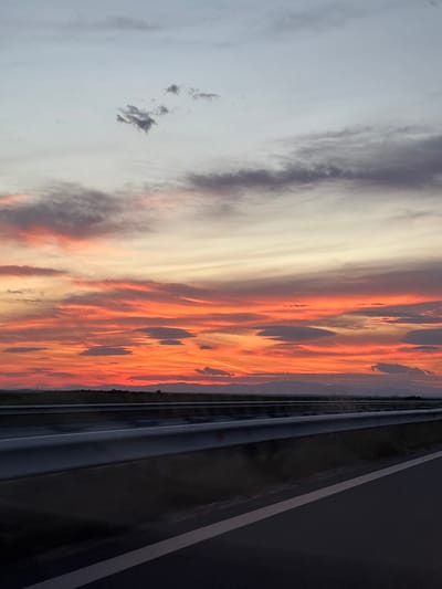 The image shows a sunset sky with orange and white clouds, with a highway in the foreground. The location is possibly near Градина, Bulgaria, based on the prompt. The highway has a guardrail, and the sky transitions from light gray to vibrant orange as the sun sets.