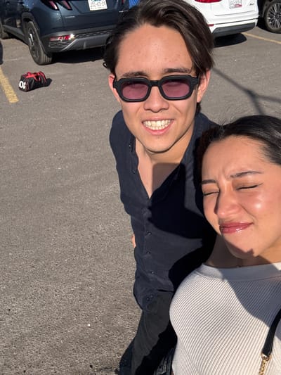 A couple poses for a selfie outdoors on asphalt. The woman, wearing a ribbed white top, is positioned slightly behind the man and has her eyes closed. The man wears dark sunglasses and a dark button-up shirt, smiling at the camera. Parts of three parked cars are visible along the left edge of the frame, and a red and black bag sits near one of them.