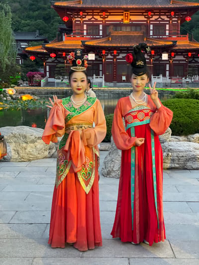 Two women in traditional Hanfu dresses pose for a photo in a park in Lintong District, China. The woman on the top wears a red Hanfu with green accents and a pearl necklace, while the woman on the bottom wears a peach-colored Hanfu with green accents and a pearl necklace. Both women are smiling and holding up peace signs. In the background, there is a traditional Chinese building with red lanterns, a pond with lily pads, and various plants and rocks. The ground is paved with gray bricks.