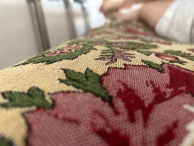 This is a close-up, low-angle shot of a floral-patterned fabric, likely upholstery on a bench or sofa. The fabric features a repeating pattern of stylized red flowers with green leaves against a pale yellow background. The texture of the woven fabric is highly detailed and in focus. In the blurred background, the lower legs of a person wearing white shorts are visible, suggesting they are sitting on the fabric. The environment appears to be indoors, possibly a home, with hints of vertical structures in the far, out-of-focus background that could be furniture legs or part of a room's architecture. There are no identifiable landmarks or indications of the specific location within Kyzyl, Russia.