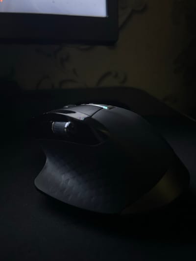 The scene depicts a dark-colored ergonomic computer mouse positioned on a black surface, identified as an indoor location in Navoiy City, Uzbekistan. The mouse presents a metallic scroll wheel, several control buttons, and a textured left-side grip. Faint green and blue indicator lights are visible on its upper casing, and a subtle golden reflection appears on its lower right rear. In the blurred background, the bottom portion of an illuminated screen or monitor is present. Behind this, a wall displays a repeating, dark floral or scroll pattern consistent with wallpaper. The ambient lighting is low, with directed light highlighting the mouse's form.