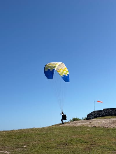 A paraglider is piloting his glider in the skies of Benoa, Indonesia. The glider is a patchwork of blue, yellow, and white. The windsock, indicating direction and speed, is fully inflated. The grassy cliff provides an edge for takeoff.