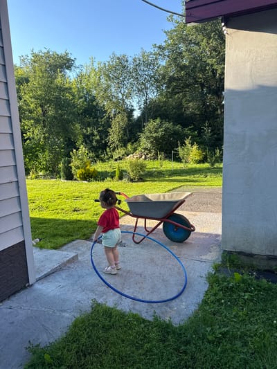 A young child, seen from behind, stands on a concrete patio near a white-sided building in Bolshie Ozerski, Russia. The child, wearing a red t-shirt and light-colored shorts, holds a blue hoop. A red and blue wheelbarrow is situated to the right of the child, positioned on the patio. The background features a lush green lawn, a line of trees, and a stone wall. The sky is clear and blue.