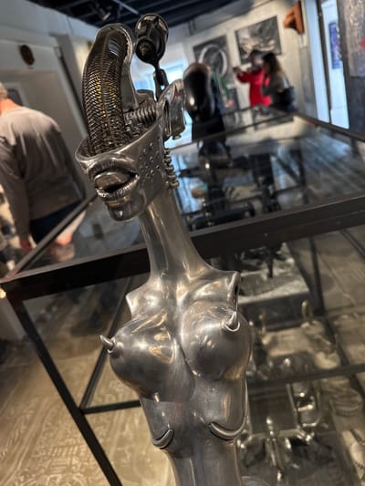 A close-up shot depicts a metallic, possibly aluminum or silver, sculpture of an elongated avian or mythological creature within a glass display case. The head of the sculpture is smooth and somewhat abstract. The body section features an intricate mesh or grill design attached to a complex mechanical apparatus. The sculpture sits within a black-framed glass display case. In the background, other display cases and items are visible, some containing what appear to be photographs or documents. Further back, through the display case, a blurred figure of a person wearing a gray shirt can be seen. The environment suggests a museum or an exhibit. The location, Epagny, Switzerland, is based on provided information.