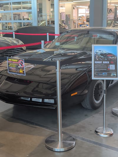 The image depicts a black 1982 Pontiac Firebird Trans Am, famously known as KITT from the television series "Knight Rider," displayed indoors, likely at an event or exhibition. The car is shiny black, with its distinctive red scanner light visible on the front. A framed poster with an image of the KITT car and the text "KNIGHT RIDER" is displayed to the right of the vehicle. To the left of the car, a yellow and black sign reads "NUR SCHAUEN - NICHT ANFASSEN!" which translates to "Look Only - Do Not Touch!". This sign also includes text about parental responsibility and legal consequences for damage, along with images of a camera and a warning symbol. The car is roped off with a red ribbon held by silver stanchions. In the background, a dimly lit industrial or workshop-like environment is visible through large glass doors and windows, suggesting a location in Böblingen, Germany, potentially at a car show or dealership. Other vehicles, some covered, are partially visible in the background. The overall lighting is artificial and somewhat dim.