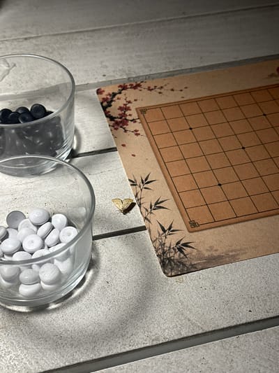 This forensic image, captured in the city of Bartula, Montenegro, displays an indoor arrangement on a light-colored, textured surface, likely composed of horizontal planks. In the left foreground, two transparent glass containers are present. The upper container holds numerous black, uniform, disc-shaped game pieces. The lower container, positioned slightly forward and to the left of the upper one, contains multiple white, disc-shaped game pieces, which exhibit subtle concentric ring patterns on their surfaces, suggesting a wooden composition.
To the right of the glass containers, a rectangular game board, appearing to be printed on paper or a thin board, is laid flat. This board features a black 9x9 grid on a light brown background. Decorative illustrations adorn its perimeter, including red cherry blossoms (sakura) along the top and upper left edge, and black bamboo stalks along the bottom and lower left edge. Two black 'star points' are visible on the grid lines.
A small, light-yellow colored moth or butterfly is resting on the textured surface, positioned between the lower glass container and the game board. The overall lighting is indirect and casts subtle shadows, particularly beneath the glass containers. No human subjects are present, and no actions are actively depicted within the scene.