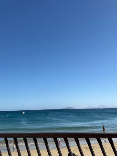 The image depicts a sunny day at Playa Blanca, Spain. The right side of the view is partially obscured by a wooden railing. Beyond the railing, a sandy beach meets the blue ocean. Several people are on the beach and in the water. A sailboat is visible in the distance. The sky is clear and blue.