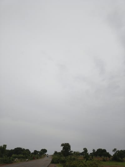 An overcast sky dominates the upper portion of the image, with subtle cloud formations visible. The lower portion of the image depicts a rural landscape with a paved road winding through it. Trees and lush green vegetation line both sides of the road. A dark-colored car is driving away from the viewer on the road, heading uphill. The scene appears to be a typical view along a road in the countryside.