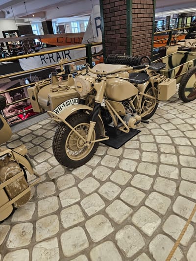 The image shows a vintage motorcycle with a sidecar, painted in a sand color. The motorcycle's license plate reads "WH-140103". The scene appears to be inside a museum, possibly in the city of Архангельское, Russia, given the request context. The motorcycle is on a stone-paved floor. In the background, elements of other exhibits are visible, including parts of a biplane labeled "R-RAR." The environment suggests an aviation and/or military history museum.