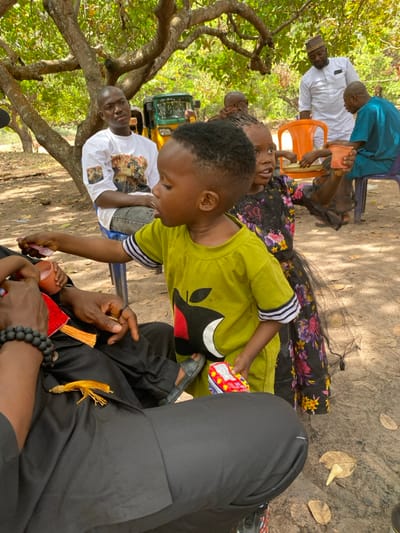 The image captures an outdoor gathering in Ochadam, Nigeria, under the canopy of large trees with exposed roots. The ground consists of dry, light brown earth scattered with fallen leaves. In the foreground, a young boy with short, dark hair, wearing a light green t-shirt featuring a black apple logo with a red bite and white outline, stands in black slide sandals. He extends his right hand, holding a small pink object, towards the lap of a partially visible seated adult whose left wrist displays a black beaded bracelet. The boy holds a red and white packaged item in his left hand. To the boy's right, a young girl, also with dark hair, wears a black dress adorned with a colorful floral pattern and a sheer black tutu-like layer. She holds a small, light orange cup in her right hand and looks towards the right side of the frame. In the midground, a bald adult male, dressed in a white t-shirt with a graphic print and blue jeans, is seated on a blue plastic chair, looking right. Behind him, a yellow and green three-wheeled auto-rickshaw is partially visible. Further right, a standing adult male in a white traditional outfit and a patterned cap observes the scene, near another seated adult male in a light blue traditional garment on a purple plastic chair. Bright daylight illuminates the entire scene.