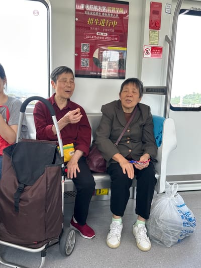The image captures a scene inside a public transit vehicle, likely a train or subway car, in Tiaodeng, China. Two elderly women are seated, occupying a row of seats, with their legs extended across the aisle. One woman is wearing a brown jacket, black pants, and white sneakers, while the other is wearing a red shirt, black pants, and maroon sneakers. There is a shopping cart near the woman in the red shirt. There is a white plastic bag on the seat beside the woman in the brown jacket. An advertisement for Chongqing Open University Jiangjin College is visible. Emergency equipment is also visible on the wall.