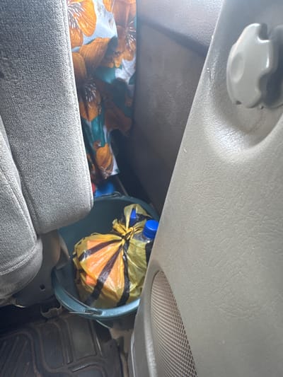 This image depicts the interior of a vehicle, likely a car or van, in Orji, Nigeria. In the foreground, a textured grey fabric seat is visible on the left. To the right, a beige-colored interior panel of the vehicle is present, featuring a circular speaker grill. Between the seat and the panel, a green bucket contains a crumpled yellow plastic bag tied with black bands, with two blue-capped bottles protruding from it. Behind the bucket and bag, a piece of fabric with an orange and teal floral pattern is draped. The floor of the vehicle, covered with a black rubber mat, is visible at the bottom of the image. The lighting suggests daytime.