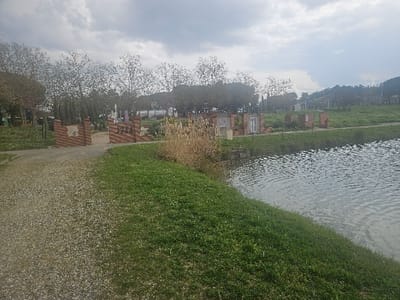 This image depicts an outdoor scene in Avignonet-Lauragais, France, under an overcast sky. The foreground on the left consists of a light-colored gravel path. To its right, a vibrant green grassy bank slopes down to a canal, whose surface exhibits ripples.
In the mid-ground, a prominent brick-built archway is visible. The left side of this archway features a crest-like symbol integrated into its brickwork, while the right side displays the letters "JAI". To the right of the archway, along the canal bank, several small, individual brick structures are arranged. These structures resemble miniature building facades, some including distinct door-like or window-like openings. Patches of dry, tall reeds line the canal's edge in this area.
The background is populated by numerous deciduous trees with minimal foliage, suggesting a period outside of full summer. Distant green hills and additional vegetation are visible under the diffused light of the cloudy sky. No individuals or specific activities are present within the scene.