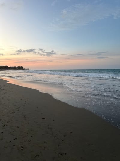 This image shows a beach scene in Cabarete, Dominican Republic. The sky is blue with some light clouds, transitioning to pink and yellow hues near the horizon. The ocean is a grey-blue color with visible waves approaching the shore. The beach is sandy and appears dark, possibly due to the time of day or wetness. There are footprints visible in the sand. Some dark shapes that look like trees are visible in the distance.