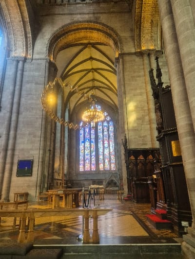 The image displays the interior of Hereford Cathedral in Hereford, United Kingdom, viewed looking towards the east end. Massive grey stone columns with ribbed detailing support high, rounded arches and a vaulted ceiling with illuminated ribs. A prominent, modern, golden, zigzagging "Chain of Light" sculpture hangs from the vaulting in the mid-ground on the left. Centrally, a large, multi-tiered chandelier is suspended before a tall, vibrant stained-glass window that dominates the rear wall, featuring predominantly blue, red, and yellow hues. To the right, a tall, dark, intricately carved wooden structure, consistent with choir stalls or a bishop's throne, exhibits Gothic spires and detailed paneling, fronted by steps covered in red carpeting. In the left foreground and mid-ground, several light-colored wooden tables and benches are arranged on a geometrically patterned tiled floor. Two silver stanchions with a black rope delineate a pathway among the tables. An abstract blue artwork with a white cross-like element is mounted on the left stone wall. The scene is devoid of people.