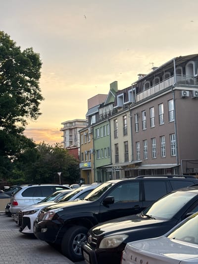 The image captures a street scene in Khamkhi, Russia, during a soft sunset. The foreground is dominated by a row of parked cars, including a black SUV, a white SUV, and a white sedan. Beyond the vehicles, several multi-story buildings line the street, showcasing a mix of architectural styles and colors, with balconies and windows visible. Trees flank the left side of the image, and the sky is a pale yellow and orange, with a few birds in flight. The overall impression is one of a quiet evening in a residential or commercial urban area.