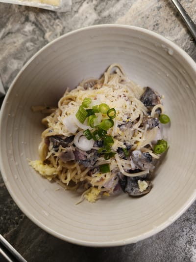 A close-up, overhead shot shows a creamy pasta dish with mushrooms and shredded cheese in a light-colored, shallow bowl. The pasta is spaghetti, tossed in a light sauce. Sliced green onions are sprinkled on top as a garnish. The bowl sits on a dark, textured countertop. The image does not contain any information about the location of Usovo-Tupik, Russia.