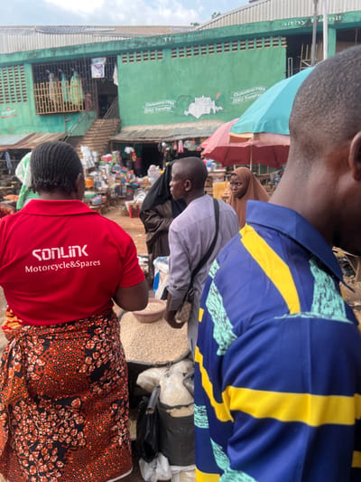 The image shows a bustling market scene in Ondo, Nigeria. The location is near the intersection of Igbatoro Street and Oyemekun Road, in the heart of the city. The image captures a diverse group of people in the market, with a focus on a woman wearing a red shirt with the inscription "SONLINK Motorcycle & Spares." She is standing near a pile of white grains, likely beans, contained in a large bowl. Her attention is directed towards the grain. Behind her is a young man in a grey shirt. He is looking towards the camera, carrying a black bag on his left shoulder. Further back, a woman is wearing a black hijab and holding a small brown bag in her right hand. To the right of the image, the back of a man wearing a blue and yellow striped shirt is facing the camera. The image shows a vibrant atmosphere, with numerous individuals interacting, and various goods being sold. The background showcases a green-colored building with a large green awning, as well as several shops and a staircase leading up to the second level. There are numerous other people, objects, and goods being sold in the market, suggesting a bustling commercial environment. The overall ambiance is a mix of everyday life and commerce.