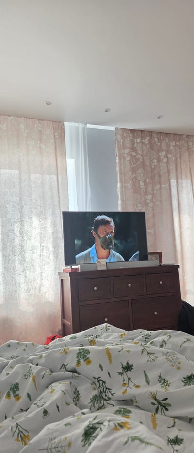 A man wearing a gas mask is visible on a television screen in a bedroom. The television is placed on top of a dark wood dresser with several drawers. The room has light-colored curtains with a floral pattern, and a white comforter with a botanical print is in the foreground. The ceiling has recessed lighting.