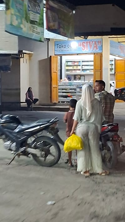 The image depicts a street scene in Keudegeudong, Indonesia. In the foreground, a black motorcycle is parked. To the right of the motorcycle, a woman wearing a light-colored headscarf and loose-fitting pants holds a bright yellow plastic bag. She is accompanied by a young child and a man wearing a plaid shirt. The group appears to be walking away from a storefront. The storefront, labeled "Toko ol "SIVA"", has shelves stocked with various items visible through the open doors. Further to the left, a woman is seated on the ground, partially obscured by shadow. The overall setting suggests a commercial area at dusk or nighttime, with artificial lighting illuminating the scene.