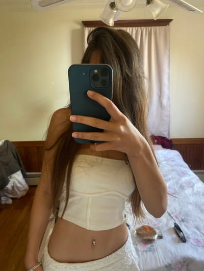 A young woman is standing in a bedroom, taking a mirror selfie. She is wearing a white strapless top and white skirt, and has a belly button piercing. She is holding a dark blue smartphone in her right hand, covering her face. The room has light yellow walls, a ceiling fan, and a wooden floor. A bed with a floral patterned comforter is visible in the background, along with a closet and a window with light-colored curtains. There is also a piece of packaged food on the bed. The location is Oak Bluffs, United States of America.