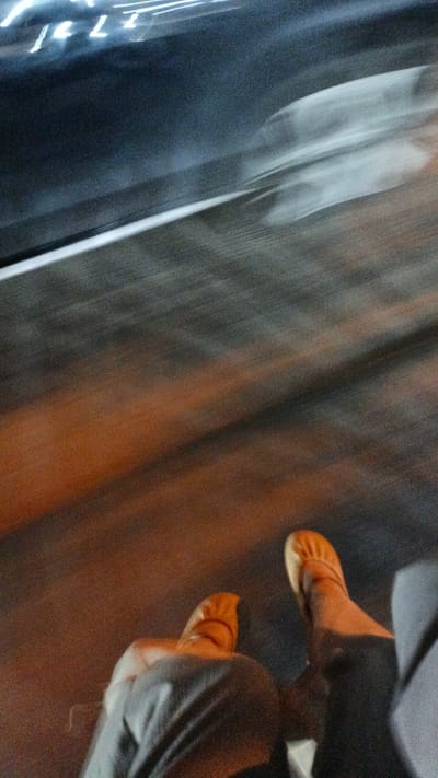 In Palohlada, Indonesia, an individual's feet, clad in light brown ballet flats, are visible. They are seated, with their legs crossed at the ankles. The person is wearing dark grey trousers. The background is blurred due to motion, suggesting movement through the city. Streaks of light in white and orange hues indicate the presence of streetlights or vehicle lights, common in urban nightscapes. The ground beneath appears to be a paved surface, possibly asphalt or concrete, with warm orange light reflecting off it. The overall impression is a snapshot of motion within an Indonesian city at night.
