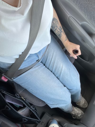 A person is seated in the passenger seat of a vehicle, wearing a white top and light blue jeans. A gray seatbelt is fastened across their body, with the buckle mechanism visible on the left. Their right arm rests on the armrest of the car door, and they are holding a dark-colored bottle with their right hand. A tattoo is visible on their left forearm. In the foreground, a black handbag is placed on the floor. The background shows the interior of the car, including the car door and seating upholstery. The overall scene suggests the person is a passenger in a car, possibly in transit. This image was taken in Bolshaya Rechka, Russia.