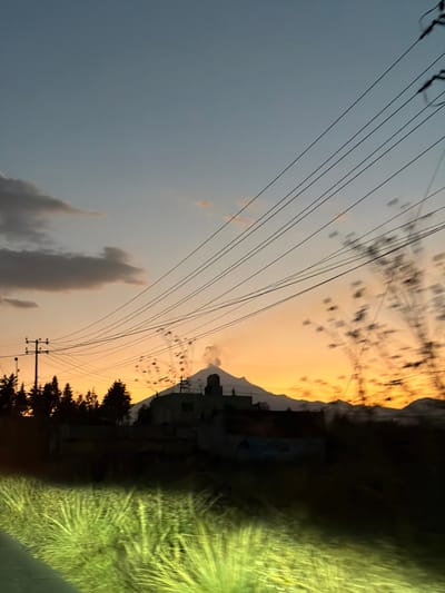 A low-angle shot captures a landscape at dusk, featuring power lines crisscrossing the sky. Mountains are silhouetted against an orange-yellow sky, while trees line the base of the mountains. Clouds punctuate the blue-gray section of the sky. A structure resembling a church stands atop a hill in the middle distance. Green foliage fills the right side of the frame. The horizon is tilted, creating an off-kilter perspective.