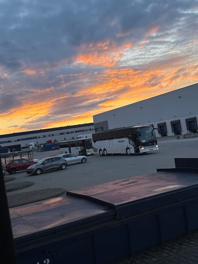 The image depicts a bus parking lot in Bielany Wrocławskie, Poland, with a large industrial building in the background. The sky is filled with dramatic, colorful clouds during what appears to be sunset. Several vehicles are visible, including a white bus with "POLBUS" branding, a silver car, and a red car. The ground is paved with interlocking stones. The overall scene suggests a quiet evening at a logistics or transportation hub.