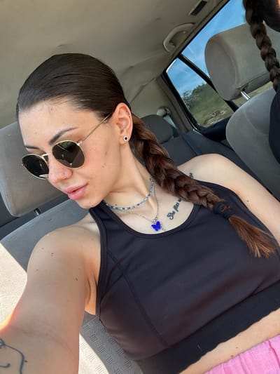 The image shows a person, likely female, in a reclined position inside a car, captured from a slightly upward angle. The individual has dark brown hair styled in a long braid that rests across their chest. They are wearing a black tank top or sports bra.
The person is wearing round, dark-tinted sunglasses. Accessories include a black stud earring and a silver hoop or bar piercing in the upper cartilage of their left ear, as well as a small silver hoop nose piercing. They have two necklaces: a multicolored beaded chain (featuring blue, green, white, and silver beads) and a silver chain with a distinct blue butterfly charm.
Several tattoos are visible: script on the upper chest/shoulder area that partially reads "She flies w", and a partial design on the lower right arm/wrist.
The setting is a car interior with gray upholstered seats and headrests. A sunroof above reveals a clear, bright blue sky with faint clouds, suggesting daytime. Through the sunroof, some greenery, likely trees, can be seen in the distance. The presence of sunlight casting shadows within the vehicle confirms bright, clear weather. On the far right, a section of pink fabric is visible. Faintly reflected in the sunroof, the word "tears" can be seen. The overall scene conveys a relaxed moment during a daytime drive in what is identified as Belopoptsi, Bulgaria.
