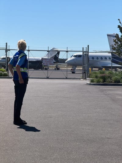 An individual wearing a blue shirt, dark pants, and a high-visibility vest stands facing away from the camera, looking towards parked airplanes. The individual's vest has "SIGNATURE AVIATION" printed on it. Behind the person is a chain-link fence with barbed wire at the top, separating the paved area from the tarmac where several small jets are parked. A "KEEP GATE CLOSED AT ALL TIMES" sign is attached to the fence. The scene appears to be an airport tarmac on a sunny day, possibly at Signature Flight Support in the Town of Southampton, United States of America.