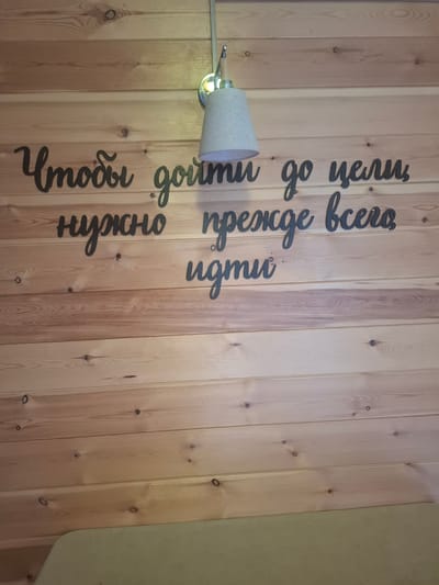 The image shows a wooden wall with black, cursive text cutouts. The text reads "Чтобы дойти до цели нужно прежде всего идти". This translates to "To reach the goal, one must first walk". Above the text, a light fixture with a conical shade is mounted on the wall. The overall setting appears to be an interior space with natural wood paneling. The location in the city of Sukhoy Ruchey, Russia, is not discernible from the image itself. There are no people or discernible actions depicted. The scene is static and focuses on the wall decoration.