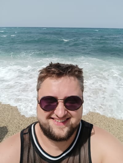 A man is taking a selfie on a beach. He is wearing sunglasses and a mesh tank top. The ocean is behind him, with waves breaking on the shore. The beach appears to be made of small pebbles or gravel. The sky is clear and bright. The location is the city of Águilas, Spain.