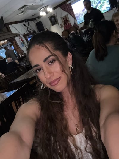 A selfie taken in an indoor dining area in Des Plaines, Illinois. The person taking the photo has long, dark, curly hair, and is wearing gold hoop earrings and a delicate gold necklace. The background shows other people, rustic decor, a television, and furniture.