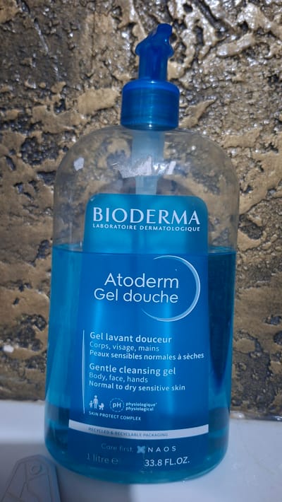 A clear plastic bottle with a blue pump dispenser is filled with blue liquid. The bottle is labeled with "BIODERMA LABORATOIRE DERMATOLOGIQUE" at the top, followed by "Atoderm Gel douche" and "Gentle cleansing gel" in English. Below that are more product details in French and English, including "Corps, visage, mains" and "Body, face, hands" and "Peaux sensibles normales à sèches" and "Normal to dry sensitive skin." There is also a "pH physiologique" symbol and "SKIN PROTECT COMPLEX." The bottom of the label shows "RECYCLED & RECYCLABLE PACKAGING," "Care first. NAOS," "1 litre e," and "33.8 FL.OZ." The bottle is set against a textured, mottled brown and beige background that resembles a wall. The image is cropped to focus on the bottle and its label. There are no people or any indication of a specific location in the city of Voinova, Moldova.
