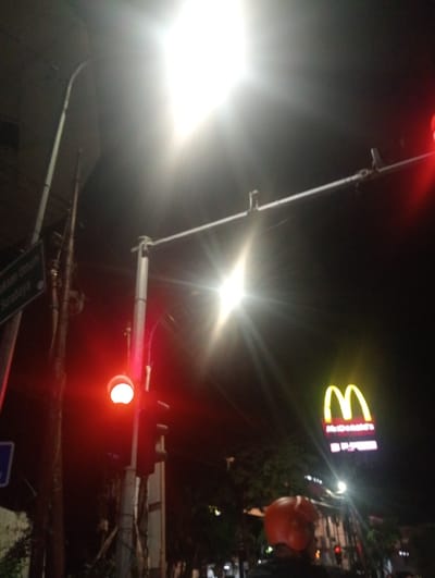 The media file depicts a night scene on a street in Surabaya, East Java, likely at an intersection. A bright street light illuminates the foreground, casting lens flares and creating a high-contrast effect. A traffic light is visible, currently displaying a red light, suggesting a stop.

In the mid-ground, a prominent McDonald's sign with its iconic golden arches is illuminated against the dark sky. Below the arches, the text "McDonald's" is legible, and further down, a pink rectangular sign with some unreadable text is also visible.

To the left of the traffic light, a street sign with green text is mounted on a pole. The visible text on this sign appears to be "Jalan Umum Surabaya," indicating a main road in Surabaya.

In the lower right portion of the frame, a person wearing an orange helmet and partially visible torso is present, possibly on a motorcycle, suggesting the presence of traffic or commuters. The overall atmosphere is urban and nocturnal, with artificial lighting dominating the scene. The time of day is clearly night, and there are no indications of weather conditions. The presence of the traffic light, street sign, and McDonald's signage firmly establishes the urban setting.