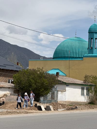 The image shows a mosque with a light blue dome and minaret in Кашат, Kyrgyzstan. The mosque is adjacent to a beige building with a blue roof. A mountain is visible in the background. In the foreground, three girls in school uniforms walk along the side of a road. The road runs beside houses.