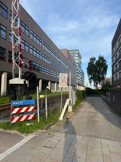 A railway crossing in Enschede, Netherlands, is captured in this image. The crossing is marked by a red and white striped barrier arm and a warning sign that reads "Wacht," indicating "Wait." A traffic signal with two black lights is also visible. The crossing is situated next to a chain-link fence that borders the railway tracks. Beyond the fence, a dark-colored building with numerous windows stretches across the background. To the right of the crossing, a paved pathway leads away, flanked by a brick wall on one side and a hedgerow on the other. A tall tree stands prominently in the mid-ground, with a building visible behind it. The sky above is clear with some wispy clouds.