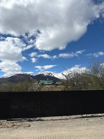 The image presents a scenic mountainous landscape under a bright blue sky, viewed through a foreground fence. The overall scene is serene and depicts natural beauty.

In the foreground, a tall, dark chain-link or mesh fence runs horizontally across the bottom of the frame, partially obstructing the view. To the right of the fence, a light-colored dirt or gravel road is visible.

Behind the fence, numerous deciduous trees, mostly bare with some emerging green foliage, suggest an early spring setting. Further in the mid-ground, a multi-story building with a prominent green roof is nestled at the base of the mountains, partially visible through the trees and above the fence line.

The background is dominated by majestic, rugged mountains, their upper slopes covered in patches of snow. Above the mountains, the sky is a vibrant blue, adorned with scattered, fluffy white clouds. The lighting is bright and even, indicative of daytime, likely mid-day.

There are no people or specific activities taking place in the image. The scene captures the tranquil and expansive environment characteristic of a natural park. No visible text is present. Given the location context, this is a view from Denali Park, United States.