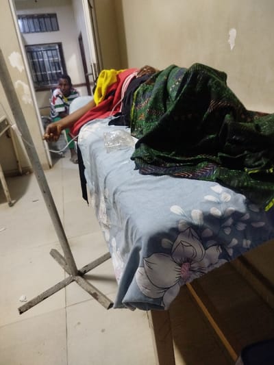 The image depicts an indoor scene within a medical facility in Mape, Nigeria. In the midground, an individual is lying on a bed covered with a light blue sheet adorned with white and purple floral patterns. The person is largely obscured by a dark green patterned blanket, with a red shirt and other multi-colored fabric (yellow, black) visible near the upper torso. The individual's right arm is extended, showing an intravenous line inserted, with tubing leading to a grey, corroded metal IV stand positioned prominently in the left foreground. A small, clear plastic wrapper rests on the bed sheet near the individual's body. In the background, an out-of-focus male figure, wearing a white t-shirt with horizontal green, black, and red stripes, is seated and looking towards the bed through an open doorway. The floor is tiled with light-colored squares, and the walls are light-toned, showing some patches of wear. Barred windows are visible in the far background.