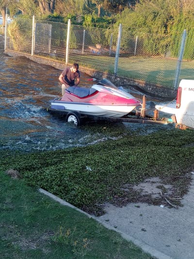 The image depicts an outdoor scene at a boat launch or canal in Villa Carlos Paz, Argentina, captured during the golden hour, indicated by the warm, low sunlight and long shadows.
In the foreground, a personal watercraft (jet ski) with red and white accents is partially submerged in a narrow waterway, likely being launched or retrieved, as its trailer wheel is also in the water. A man, wearing a dark t-shirt, is seated on the rear of the jet ski.
To the right of the water, a densely vegetated bank covered in small green plants borders the canal, transitioning into a paved or gravel path and then a patch of green grass. On the left side of the frame, a chain-link fence with metal posts runs parallel to the water. Beyond the fence, lush green trees and bushes are visible, illuminated by the warm light.
Further down the waterway, behind the jet ski, part of a larger white boat with red accents is visible, likely docked or moored. The letters "MO" are discernable on its hull. The water surface reflects the surrounding elements and the golden light. The scene conveys a calm, recreational atmosphere, characteristic of a waterfront area.