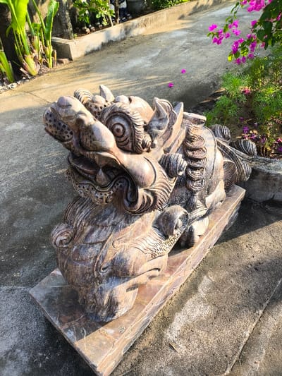 A weathered, dark reddish-brown carved stone statue, resembling a mythical Pixiu or a similar creature, is positioned outdoors on a rectangular stone plinth. The statue features intricate details, including a prominent snout, textured scales, large circular eyes, and a relief carving of stacked circular coins on its right flank. It rests on a rough, light gray concrete ground surface. To the left, large green leafy plants with thick stalks grow from a raised earth bed or planter. To the right, a bush with vibrant pink-purple flowers, possibly bougainvillea, is visible, with several detached petals scattered on the concrete. The scene is illuminated by strong natural sunlight, casting distinct shadows. No individuals are present, and no direct actions or interactions are depicted. This scene is situated within Thành phố Tuy Hòa, Vietnam.