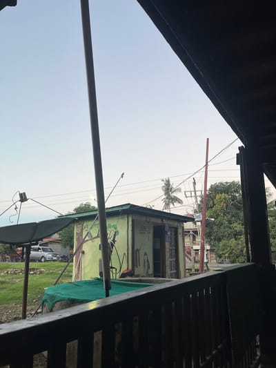 The image shows an outdoor scene in Turumame, Indonesia, at what appears to be dusk. In the foreground, a dark wooden railing with vertical slats overlooks a green tarp-covered structure, possibly a water tank or pond. Beyond this, a pale green, box-like building is visible with murals painted on its side depicting a tree with a bird and a woman. A satellite dish is positioned to the left of the building, and a tall, thin pole stands vertically in front of it.

In the background, a silver SUV is parked on a grassy area next to a road. A utility pole with wires and a transformer is also present, along with lush green trees and a palm tree. The sky is a pale blue, indicating the end of daylight. The overall impression is a casual, possibly rural or semi-urban setting in Indonesia.