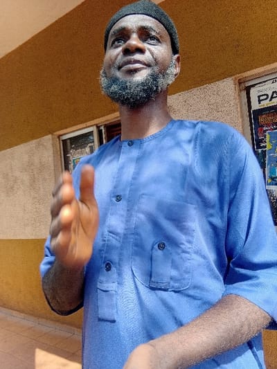 A man wearing a black skull cap and a light blue button-up shirt faces the camera with an outstretched hand. He has a graying beard. The setting appears to be outdoors with a yellow wall behind him, possibly the side of a building. A window with what seems like posters is visible.