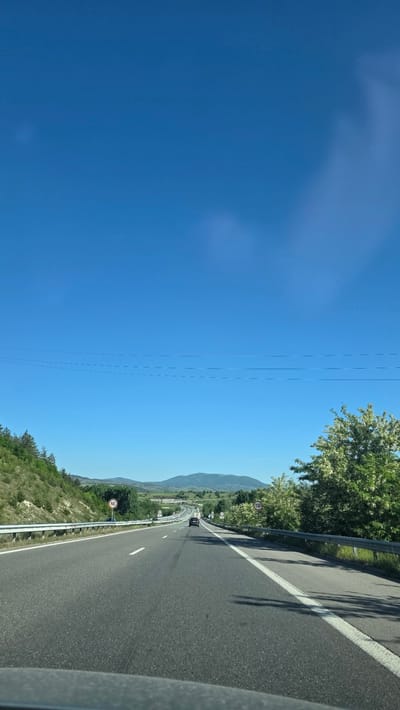 The image shows a view from a vehicle traveling on a highway near Barakovo, Bulgaria, on a sunny day. The sky is clear blue, occupying a large portion of the frame. Power lines run parallel to the highway. The highway has multiple lanes and a guardrail is visible on the side. Speed limit signs of "100" are posted. Vegetation, including trees and shrubs, flanks the road, and a mountain range is visible in the distance. Another vehicle is on the road ahead.